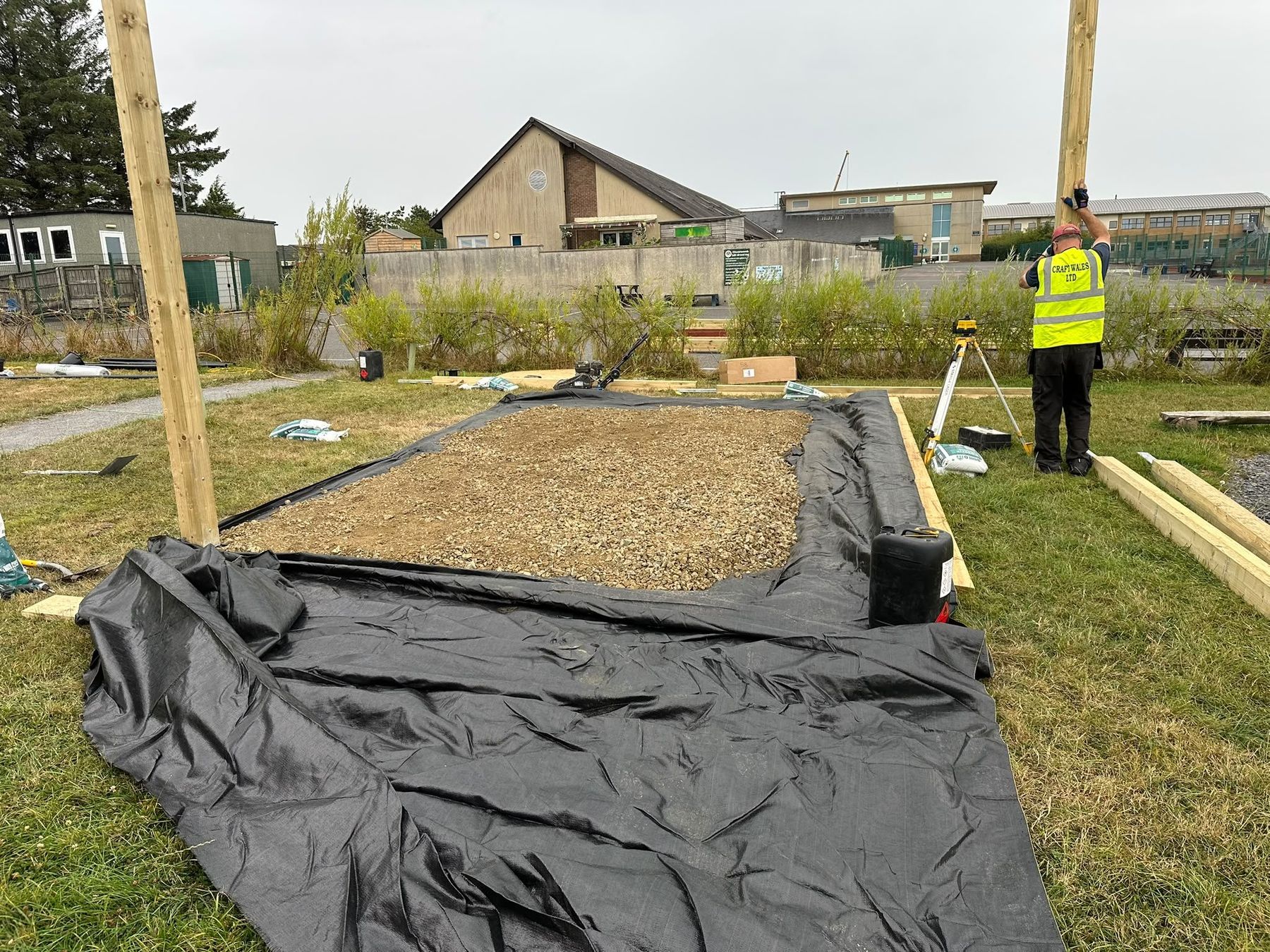 Installer in a high-visibility vest preparing the ground with membrane and gravel before a timber build