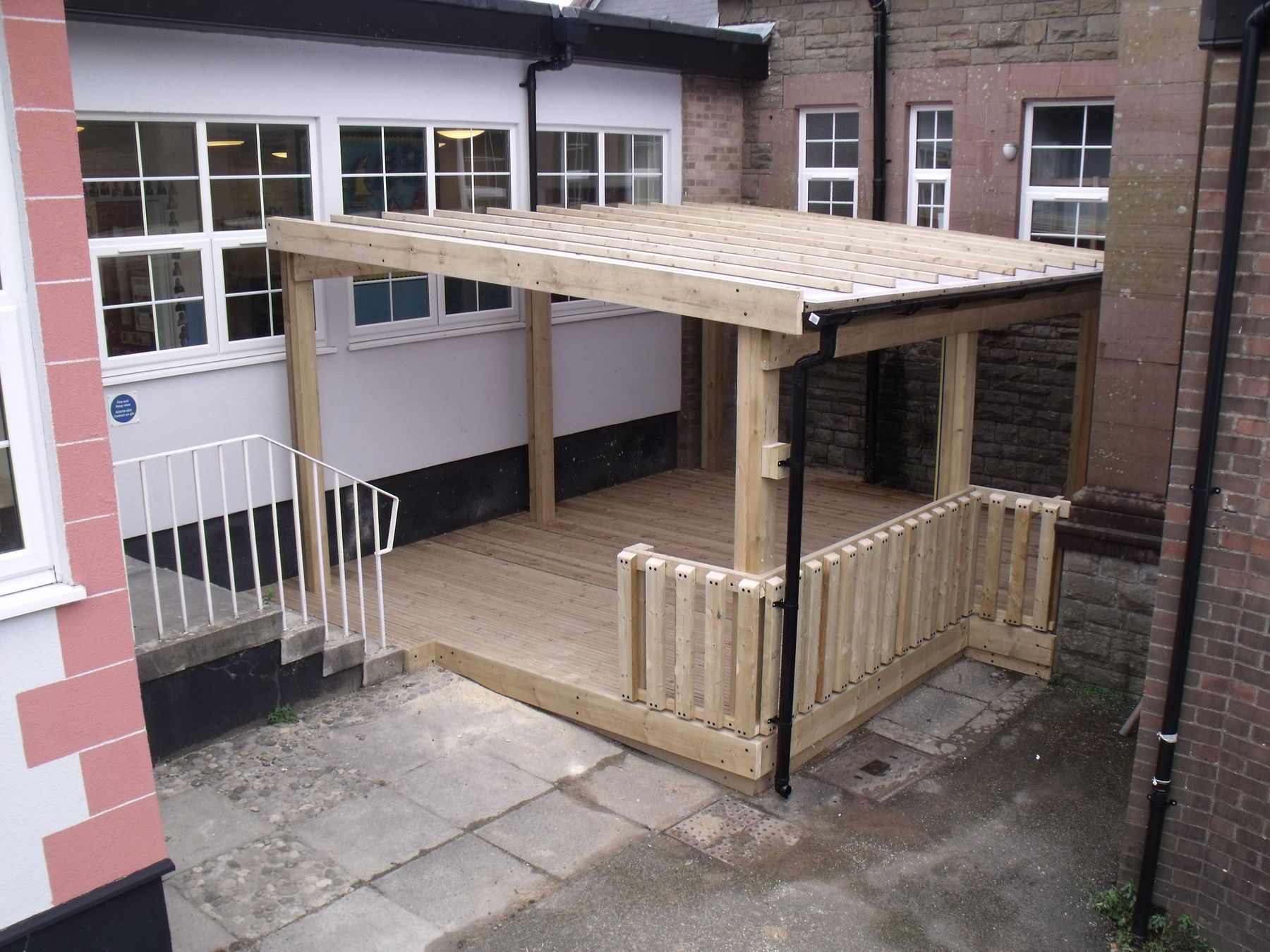 Raised timber deck with spindle balustrade and a roof canopy, built against a brick building