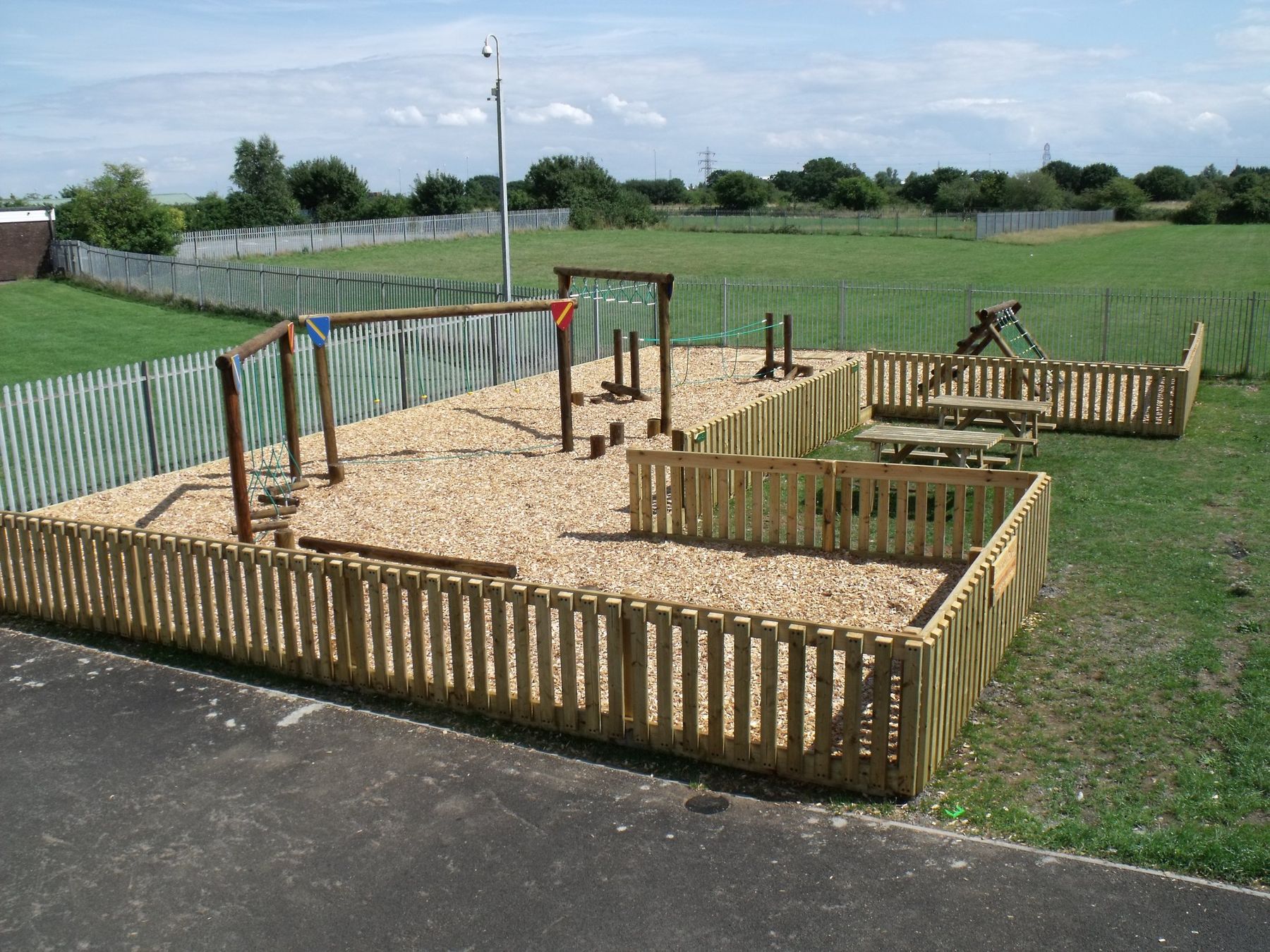 Timber trim trail with balance beams in a fenced bark-chip area beside a playing field