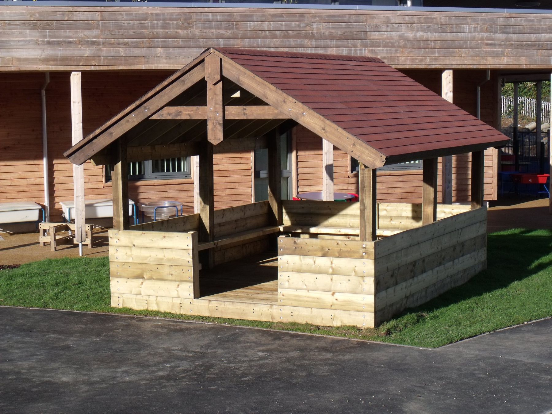 Illustrative photograph: Timber barn shelter with a red tiled roof and solid timber sides, on grass beside a school building