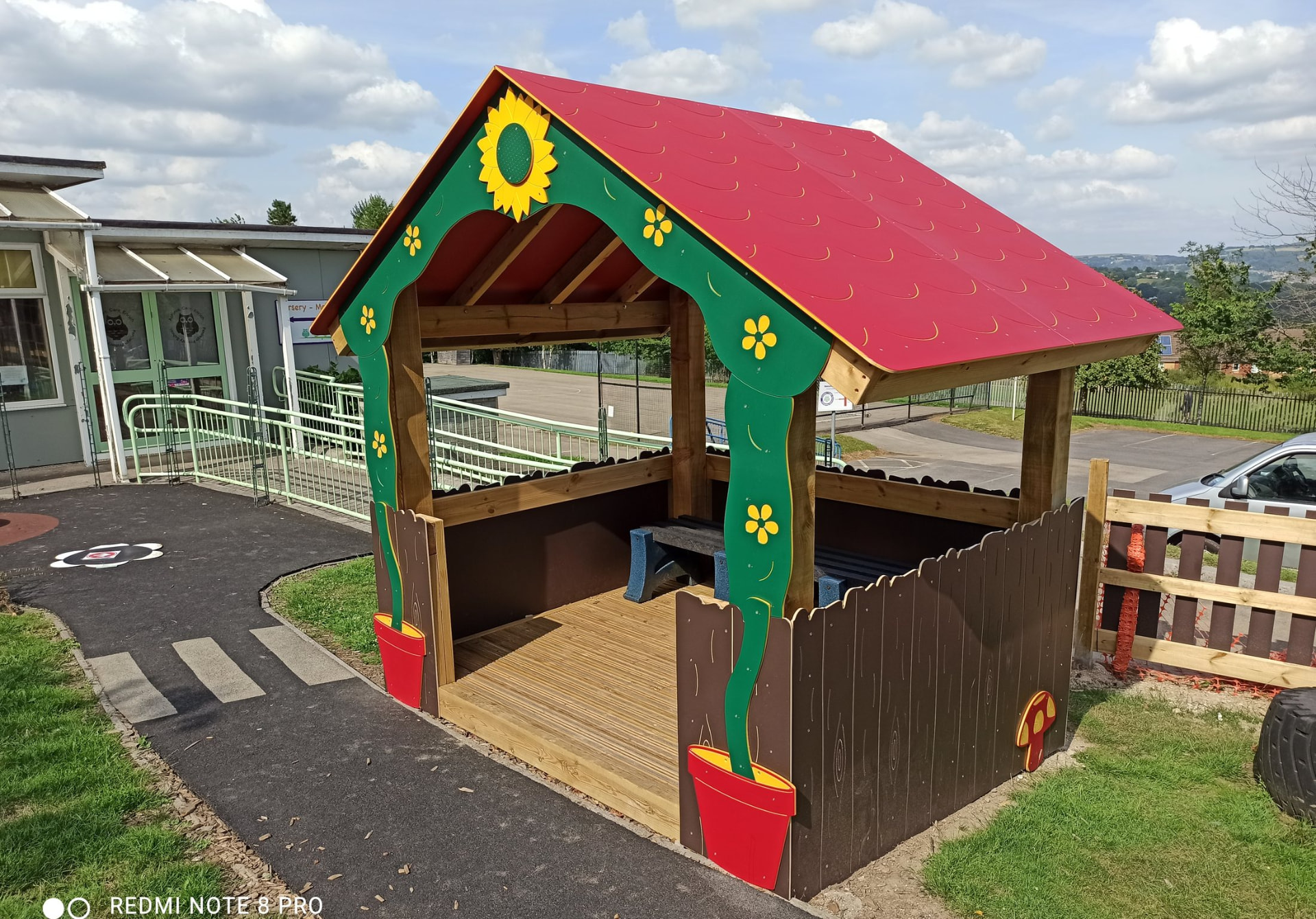 Timber play hut with a red roof and green and yellow sunflower decorations, in a school playground