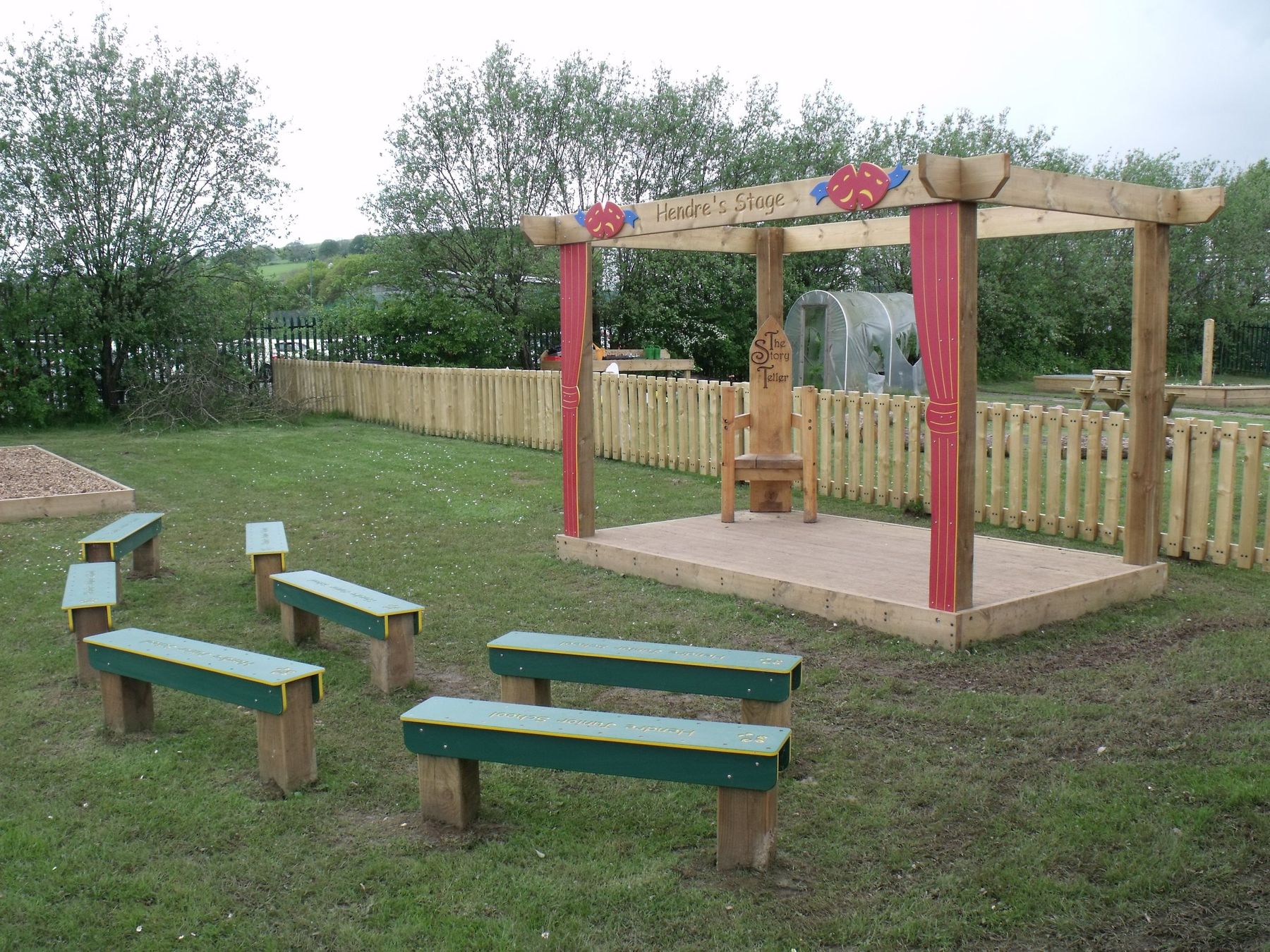 Small timber stage with a red pergola frame and green bench seating on grass