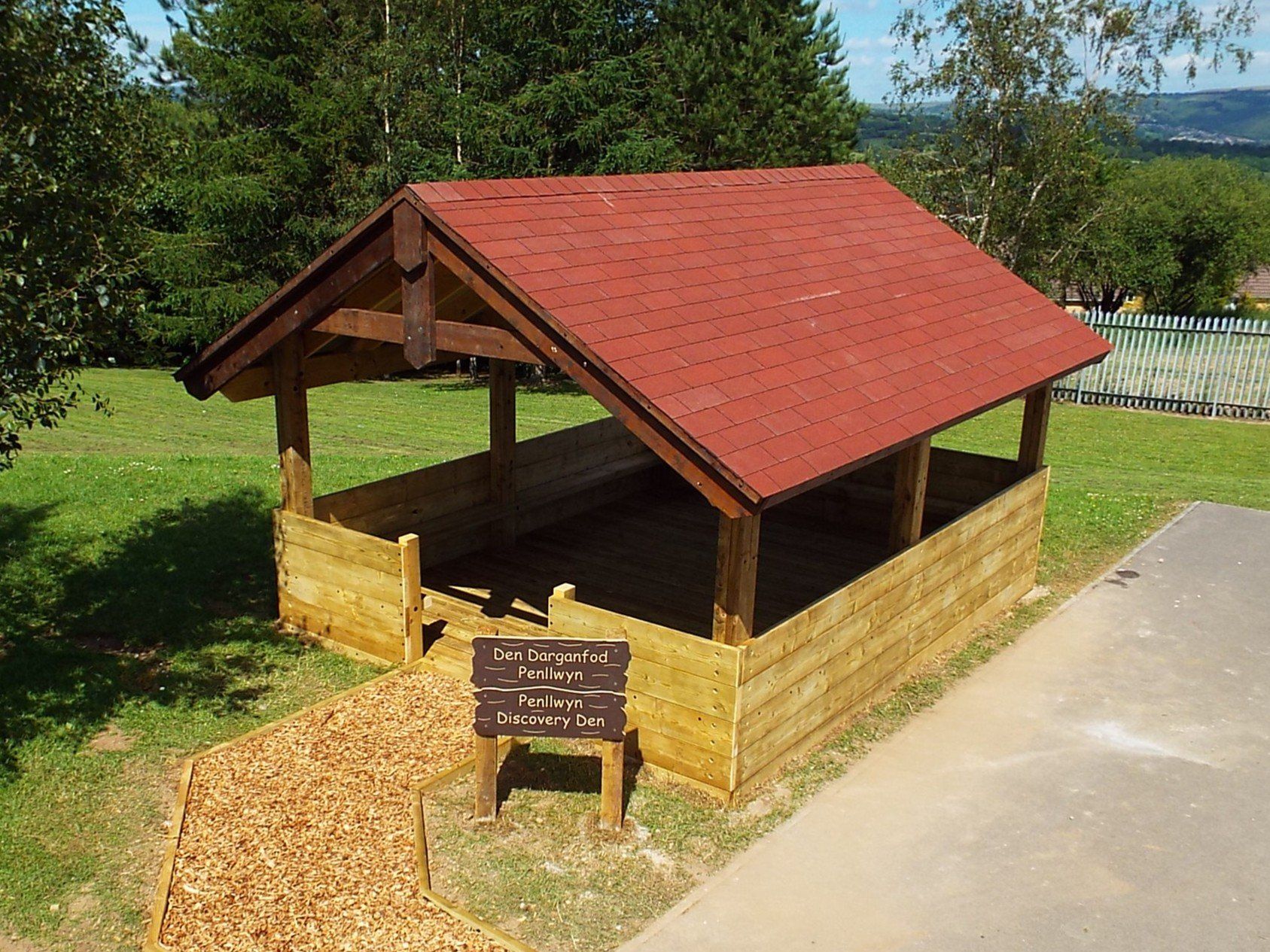 Timber barn shelter with a red roof on grass, with a carved timber sign at its entrance