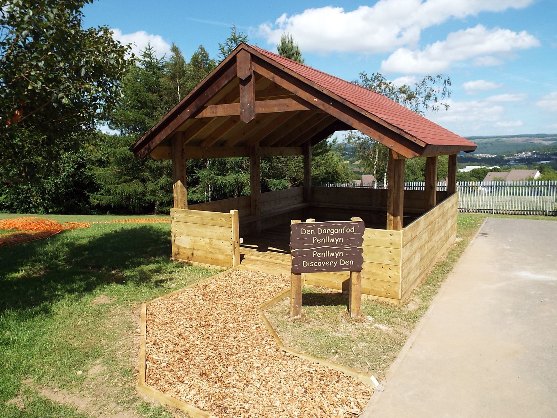 Timber barn shelter with a red roof and carved sign, beside a bark path and a school field