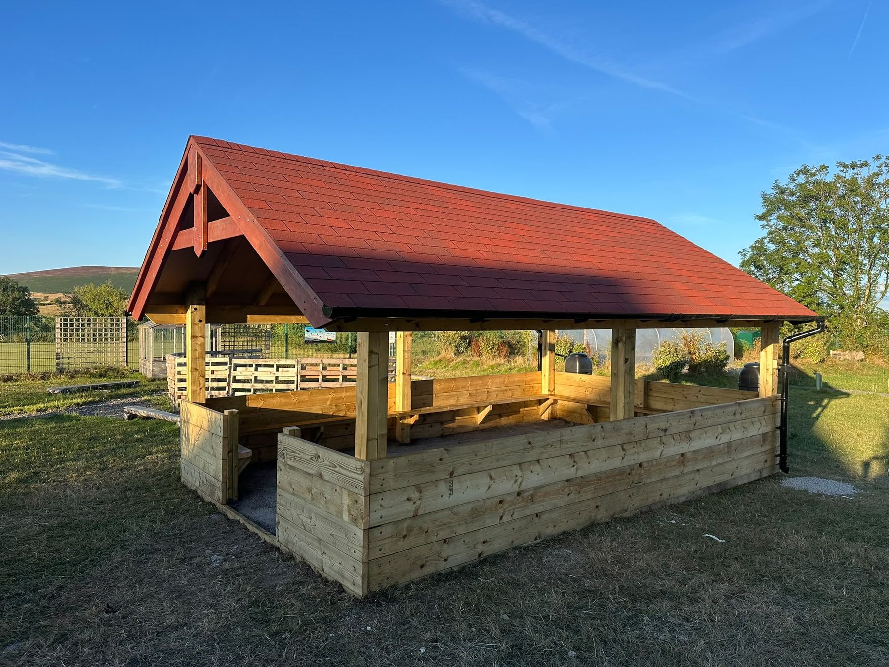 Barn-style timber shelter with a red tiled roof and boarded lower walls, standing on grass under a blue sky