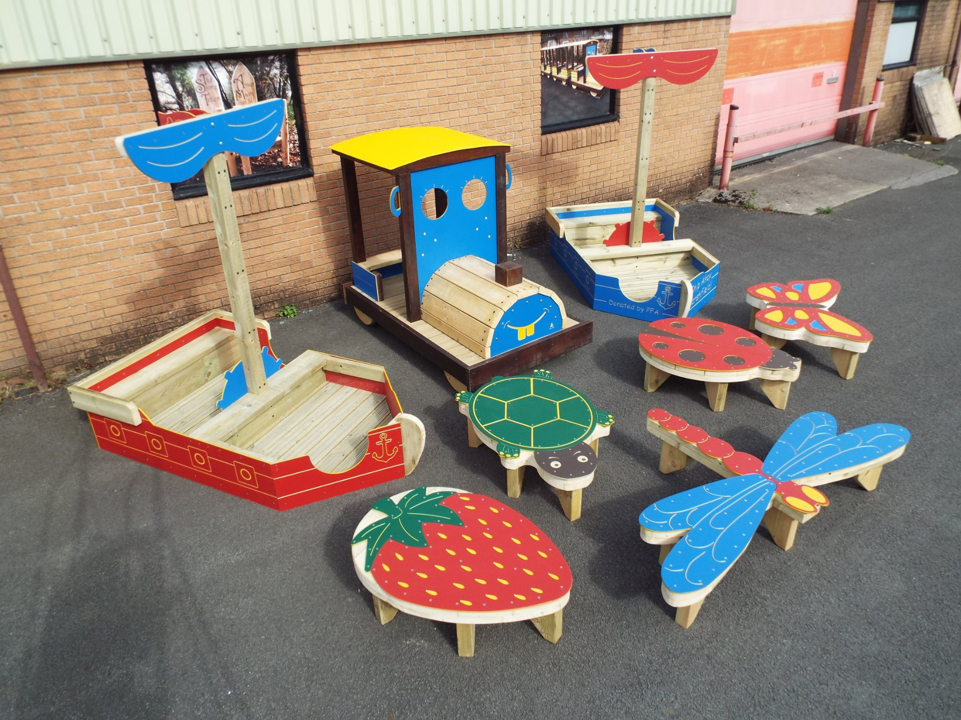 Colourful timber play tables and sandpits shaped as a ladybird, a turtle, a butterfly, a dragonfly and a strawberry, photographed outside the workshop